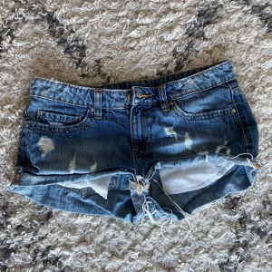 Korta lågmidjade jeansshorts - Snygga blå jeansshorts med slitna detaljer och fransig kant. Klassisk femficksmodell med låg midja. Skriv gärna ett meddelande för bild på eller vid fråga! 