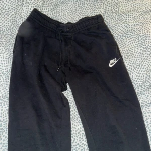 Svarta mjukisbyxor från Nike - Svarta mjukisbyxor från Nike med klassisk logga på låret. Byxorna har dragsko i midjan för skön passform och är gjorda i mjukt bomullsmaterial. Perfekta för chill eller träning. Snygg och enkel design som funkar till det mesta.