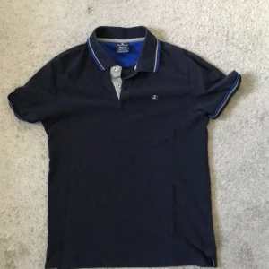 Mörkblå Champion pikétröja M - Snygg mörkblå pikétröja från Champion med klassisk krage och blå/vita detaljer på krage och ärmslut. Broderad Champion-logga på bröstet. Perfekt för en clean och sportig stil.