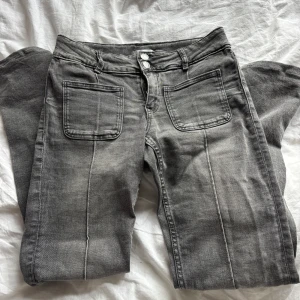 Grå bootcut jeans med raka sömmar - Snygga grå jeans med bootcut och markerade sömmar längs benen och stora fyrkantiga fickor fram. Modellen har bootcut-ben och klassiska bakfickor. Jeansen har dubbelknäppning och bälteshällor, perfekta för en cool och avslappnad look.