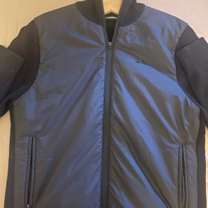 Svart bomberjacka från J.Lindeberg - Snygg svart bomberjacka från J.Lindeberg med ribbad krage och ärmslut. Jackan har dragkedja framtill, två fickor och diskret JL-logga på bröstet. Materialmix med glansig polyester på kroppen och stickade detaljer på ärmar och krage. Perfekt för en clean och stilren look.