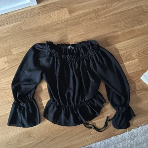 Svart offshoulder blus - Snygg svart offshoulder blus med volangdetaljer och långa ärmar. Perfekt för en stilren look. Blusen har en elastisk midja och är tillverkad i ett mjukt material.