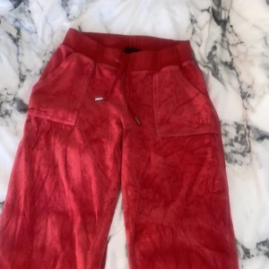 Röda mjukisbyxor med matchande huvtröja - Säljer ett set med röda mjukisbyxor och matchande huvtröja från Juicy Couture. Byxorna har snörning i midjan och bakfickor med knappar. Materialet är mjukt och bekvämt, perfekt för en avslappnad stil.