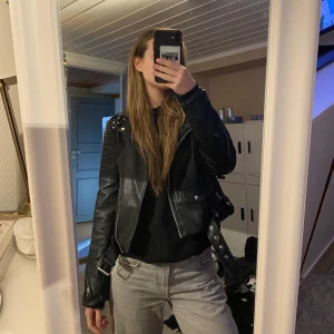 Svart skinnjacka med nitar och graffiti - Cool svart skinnjacka med nitar på axlarna och graffiti-design på ryggen. Jackan har dragkedja och bälte i midjan för en edgy look. I fint skick🫶🏼
