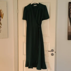 Grön klänning från Zack - Elegant grön klänning från Zack med omlott-design och knytband i midjan. Klänningen har korta ärmar och en volangdetalj vid fållen. Perfekt för en stilren look.