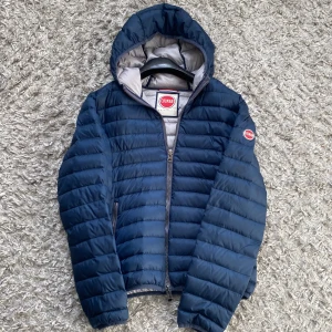 Colmar vårjacka - Snygg blå pufferjacka från Colmar med huva och dragkedja. Perfekt för kyliga dagar. Jackan har en quiltad design och en logotyp på ärmen. Storlek 48 men sitter som S - hör av dig vid frågor!