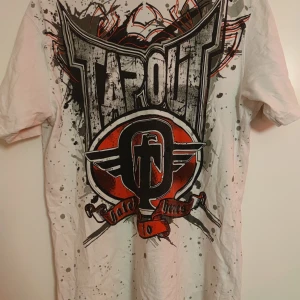 Vit T-shirt från Tapout - Säljer en cool vit T-shirt från Tapout med ett stort tryck i svart och rött på baksidan. T-shirten har korta ärmar och ett stänkmönster över hela tyget. Perfekt för dig som gillar en edgy stil! (Shoutout till ai🔥💯🙏) Mellan M och L storlek