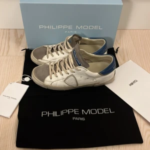 Philippe model skor - Snygga Philippe Model sneakers i vitt med blå detaljer. Skorna har en rund tå och snörning framtill. Perfekta för en stilren look med en touch av färg. Riktigt fina men använda lite små skador lite här och där pris kan diskuteras. Dom är mer slitna än vad dom ser ut att vara har bara tvättat en sko orkar inte tvätta båda därför priset också.