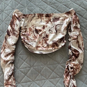 Mönstrad offshoulder topp - Säljer en unik offshoulder topp med ett abstrakt mönster i beige och bruna toner. Toppen har långa ärmar med rynkade detaljer som ger en trendig look med utsvängda armar .Perfekt för att sticka ut i mängden!
