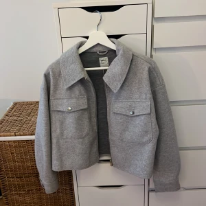 Grå croppad jacka från ONLY - Säljer en stilren grå croppad jacka från ONLY. Jackan har en klassisk krage och två stora fickor med knappar framtill. Perfekt för en trendig look!