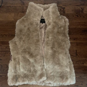Beige pälsväst från Saki - Säljer en supermjuk och fluffig beige pälsväst från Saki. Perfekt för att hålla sig varm och stilren under höst och vinter. Västen har en snygg krage och är i mycket bra skick. Passar perfekt över en tröja eller klänning för en chic look.