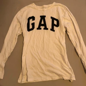 Vit långärmad tröja från GAP - Säljer en klassisk vit långärmad tröja från GAP med svart logga på bröstet. Perfekt för en avslappnad stil och enkel att matcha med andra plagg.