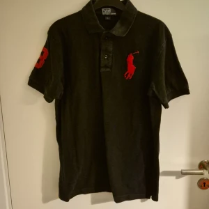 Svart pikétröja från Polo Ralph Lauren - Snygg svart pikétröja från Ralph Lauren med röd broderad logga på bröstet och siffran 3 på ärmen. Perfekt för en stilren look med klassisk krage och knappar framtill.