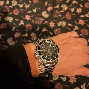 Silverfärgad klocka med svart urtavla - Säljer en stilren klocka med silverfärgad länk och svart urtavla. Klockan har tre mindre urtavlor och en tydlig minutmarkering runt ringen. Perfekt för den som gillar en klassisk look.