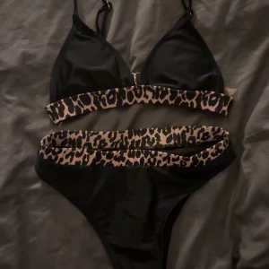 Svart bikini med leoparddetaljer - Snygg svart bikini med leopardmönstrade detaljer på både över- och underdel. Överdelen har justerbara axelband och en bekväm passform. Perfekt för en dag på stranden eller vid poolen. Aldrig använd men inga prislappar kvar