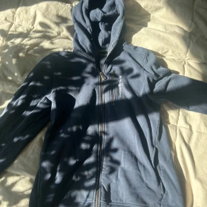 Mörkblå hoodie från Sail Racing - Snygg mörkblå hoodie från Sail Racing med dragkedja och huva. Perfekt för kyliga dagar med sin sportiga design och bekväma passform. Den har en praktisk bröstficka med dragkedja för småsaker. Stl 176