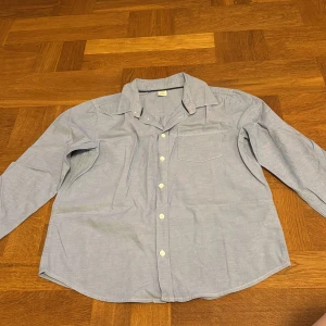 Blå skjorta - Säljer en klassisk blå skjorta från Lindex med långa ärmar och knappar framtill. Skjortan har en bröstficka och är perfekt för en stilren look. Tillverkad i ett bekvämt material.