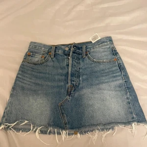 Jeanskjol från Levi's - Säljer en snygg jeanskjol från Levi's i klassisk blå denim. Kjolen har en rå kant nertill och femficksdesign med knapp och dragkedja framtill. Perfekt för en avslappnad stil.