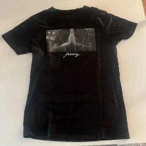 Svart t-shirt med tryck - Säljer en svart t-shirt med ett stilrent tryck av händer i bön och texten 'pray'. T-shirten har en normal passform och är kortärmad, perfekt för en avslappnad stil.
