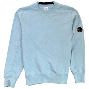 Ljusblå sweatshirt från C.P. Company - Snygg ljusblå sweatshirt från C.P. Company med rund hals och ribbade muddar. Tröjan har en cool detalj med en svart logotyp på ärmen. Perfekt för en avslappnad stil.