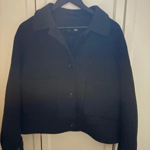 Svart kappa från lager 157 - Snygg svart kappa från lager 157 med klassisk krage och knappar framtill. Perfekt för en stilren look. Kappan har långa ärmar och två fickor framtill.