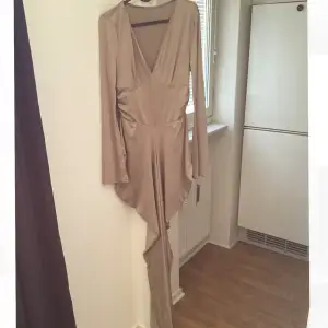 Elegant beige långklänning med djup v-ringning och långa ärmar. Klänningen har en asymmetrisk design med volangdetaljer som ger en unik look.