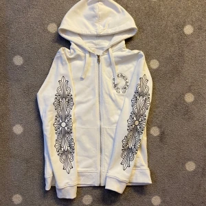 Vit hoodie från Chrome Hearts - Säljer en vit hoodie från Chrome Hearts med dragkedja och detaljerade svarta tryck på ärmarna och ryggen. Hoodien har en justerbar huva och är perfekt för en stilren look. Den har också ett unikt märke på dragkedjan.