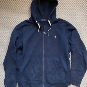 Mörkblå hoodie från Polo Ralph Lauren - Säljer en snygg mörkblå hoodie från Polo Ralph Lauren med dragkedja och klassisk logga på bröstet. Perfekt för en avslappnad stil. Den har en justerbar huva med vita snören och praktiska fickor framtill.