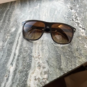 Solglasögon från Ray-Ban - Snygga bruna solglasögon från Ray-Ban med klassisk design. De har en svart båge och mörka glas som ger ett stilrent intryck. Perfekta för soliga dagar och en tidlös accessoar i garderoben.