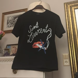 Saint laurent t-shirt  - Snygg svart t-shirt från Saint Laurent med ett coolt tryck av röda läppar och texten 'Saint Laurent' i vitt. Perfekt för en trendig look. Tröjan är extraordinär och har det lilla extra i sig, väldigt charmig och kan användas när som helst i din vardag. Fler frågor via DM ! 👇