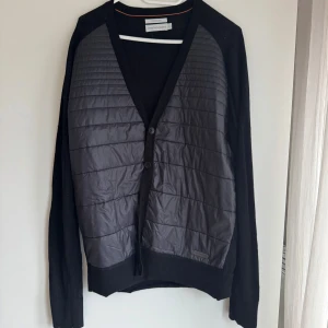 Svart kofta från Peak Performance - Snygg svart kofta från Peak Performance med quiltade detaljer framtill. Den har en v-ringad design med knappar och är tillverkad i merinoull för extra komfort. Perfekt för kyligare dagar.