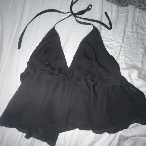 Svart halterneck topp - Snygg svart halterneck topp med knytband i nacken. Toppen har en luftig passform med volangdetaljer och är perfekt för varma dagar.
