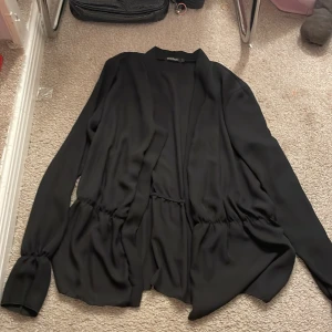 Svart cardigan jacka från Boohoo - Säljer en stilren svart cardigan jacka från Boohoo. Jackan har långa ärmar och en elegant, lös passform med knytning i midjan. Perfekt för att lägga till en touch av elegans till din outfit.