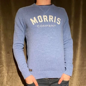 Stickad tröja från Morris - Säljer en snygg blå tröja från Morris med vit text på bröstet. Tröjan har långa ärmar och ribbade muddar vid ärmslut och nederkant. Perfekt för en stilren look.