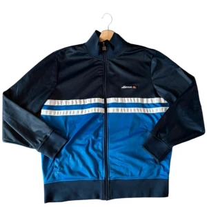  Ellesse - Snygg träningsjacka från Ellesse i blått och svart med vita ränder över bröstet. Jackan har dragkedja och ribbade muddar vid ärmslut. Perfekt för en sportig look!