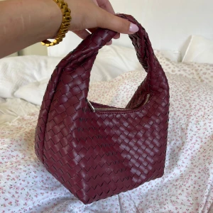 Vinröd flätad handväska från Gina Tricot - Snygg vinröd handväska från Gina Tricot med flätat mönster. Perfekt accessoar för att lyfta din outfit. Väskan har ett praktiskt handtag och är rymlig.