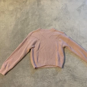 Rosa stickad tröja - Säljer en söt rosa stickad tröja med ett unikt mönster framtill. Tröjan har långa ärmar och en hög halsringning som ger en mysig känsla. Perfekt för kyligare dagar!
