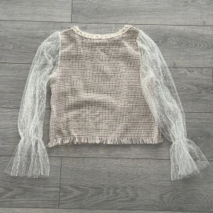 Beige topp med spetsärmar och pärldetaljer - Säljer en elegant beige topp med unika spetsärmar och pärldetaljer vid halsringningen. Toppen har en fin textur och fransar vid nederkanten som ger en chic touch. Perfekt för att lyfta vilken outfit som helst!