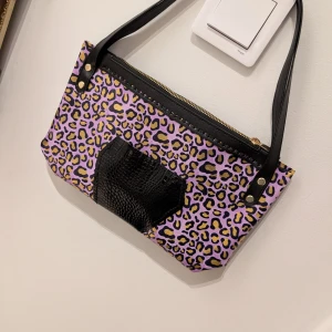 Leopardmönstrad axelväska med dragkedja - Cool axelväska med leopardmönster i lila och guld. Väskan har en svart dragkedja och svarta handtag. Den har också en svart detalj framtill som ger en extra touch. Perfekt för att ge din outfit en färgglad och djärv look. Obs! Handsydd! 