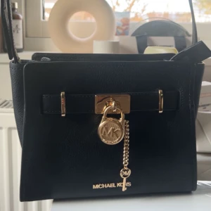 Svart väska från Michael Kors - Snygg svart handväska från Michael Kors med gulddetaljer och ett dekorativt lås framtill. Den är äkta och i ny skick! Ny pris är runt 1500-2000kr