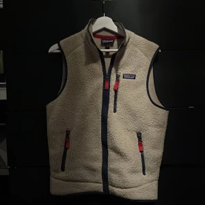 Beige fleeceväst från Patagonia - Säljer en beige fleeceväst från Patagonia med dragkedja och tre fickor med kontrasterande detaljer i rött och blått. Perfekt för lager-på-lager under kyliga dagar. Västen har en hög krage för extra värme. Perfekt plagg att ha till våren. Varan är använd men det finns inga synliga defekter förutom ullen som inte är riktigt lika ”fluffig” längre(kommer inte att synas på längre avstånd). Skick 7/10. 