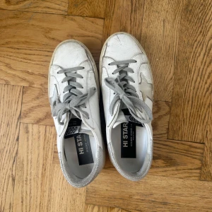 Vita sneakers från Golden Goose - Säljer ett par vita Golden Goose Hi Star sneakers med grå snörning och silverdetaljer. Skorna har en stjärna på sidan och texten 'Golden Goose' längs sulan. Tillverkade i Italien och designade för en unik look.