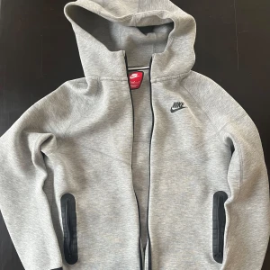 Grå hoodie Nike Tech - Säljer en grå hoodie från Nike med dragkedja och huva. Jackan har svarta detaljer och är tillverkad i ett mjukt material. Sparsamt använd, i fint skick, nästintill nyskick