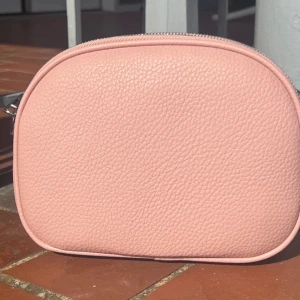 Rosa axelväska med dragkedjor - Snygg rosa axelväska i syntetmaterial med tre separata fack och dragkedjor. Perfekt för att hålla ordning på dina saker. Justerbar axelrem ingår.