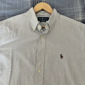 Vit och blå randig skjorta från Ralph Lauren - Snygg vit och blå randig skjorta från Ralph Lauren med klassisk krage och broderad logga på bröstet. Skjortan är helt oanvänd och säljs då den är för stor. För fler bilder, hör av dig.
