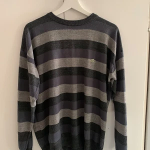 Randig tröja från Lacoste - Säljer en randig tröja från Lacoste i grå och mörkblå nyanser. Tröjan har långa ärmar och en klassisk rund halsringning. Perfekt för en stilren och avslappnad look.