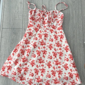 Vit och röd blommig klänning - Charmig vit klänning med röda blommor och smala axelband. Den har en söt knytning framtill och är perfekt för varma dagar. Klänningen är kort och har en lätt och luftig känsla.