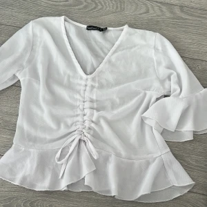 Vit blus från Boohoo - Säljer en stilren vit blus från Boohoo med v-ringning och volangdetaljer. Blusen har trekvartsärmar och ett knytband framtill som ger en snygg passform. Perfekt för en chic look!
