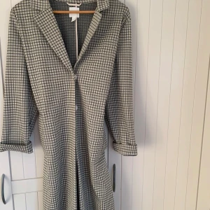 Rutig kappa - Lång kappa med klassiskt mönster i svart och beige. Kappan knappar framtill och ordentliga fickor. Perfekt för våren eller en kylig sommarkväll. 