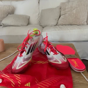 Adidas F50 fotbollsskor - Säljer ett par Adidas F50 fotbollsskor i vitt och rött med svarta detaljer. Skorna har en strömlinjeformad design med snörning och en lätt syntetisk överdel. Perfekta för snabba rörelser på planen.
